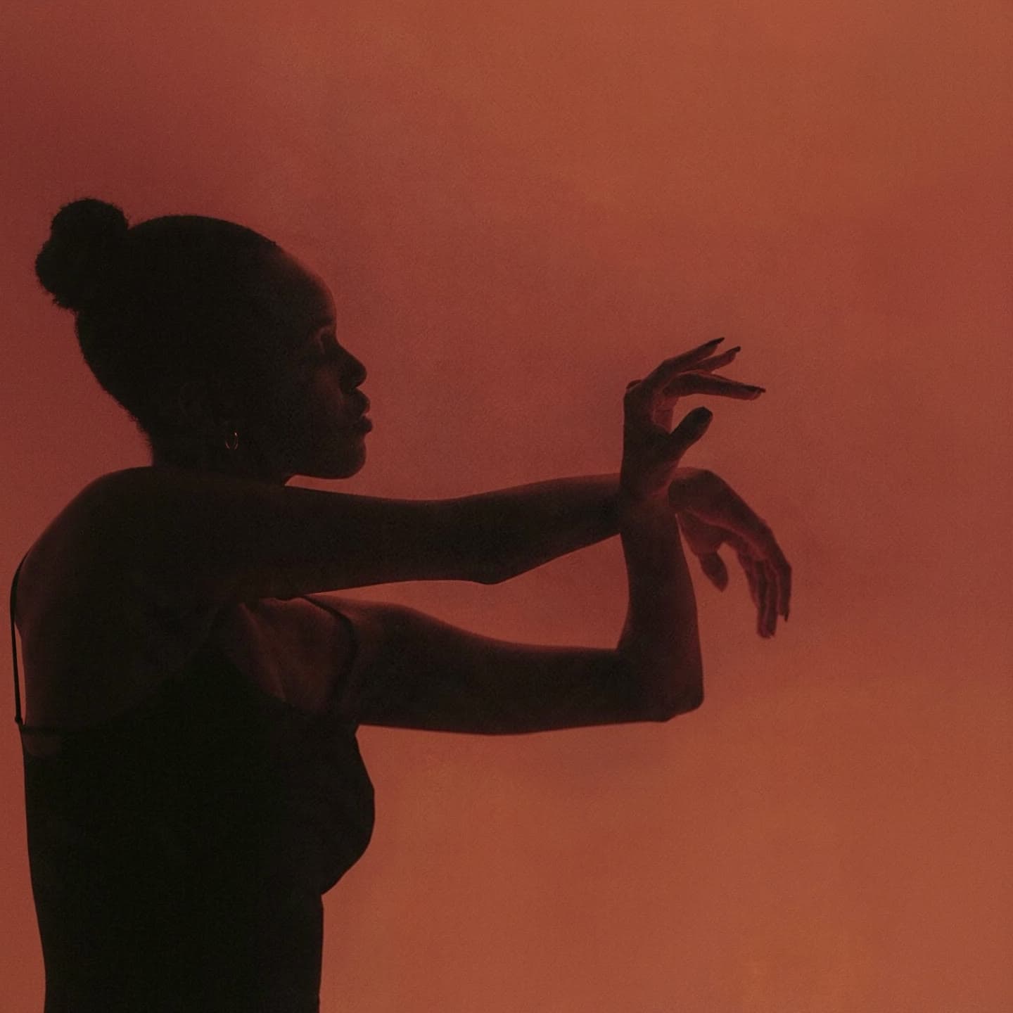 A dancer in silhouette against a warm amber backdrop, one arm drawn across the body and the hands held poised in front of her.